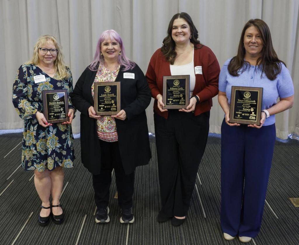 Four women posing together, each holding an award plaque, in a formal setting with a neutral backdrop.